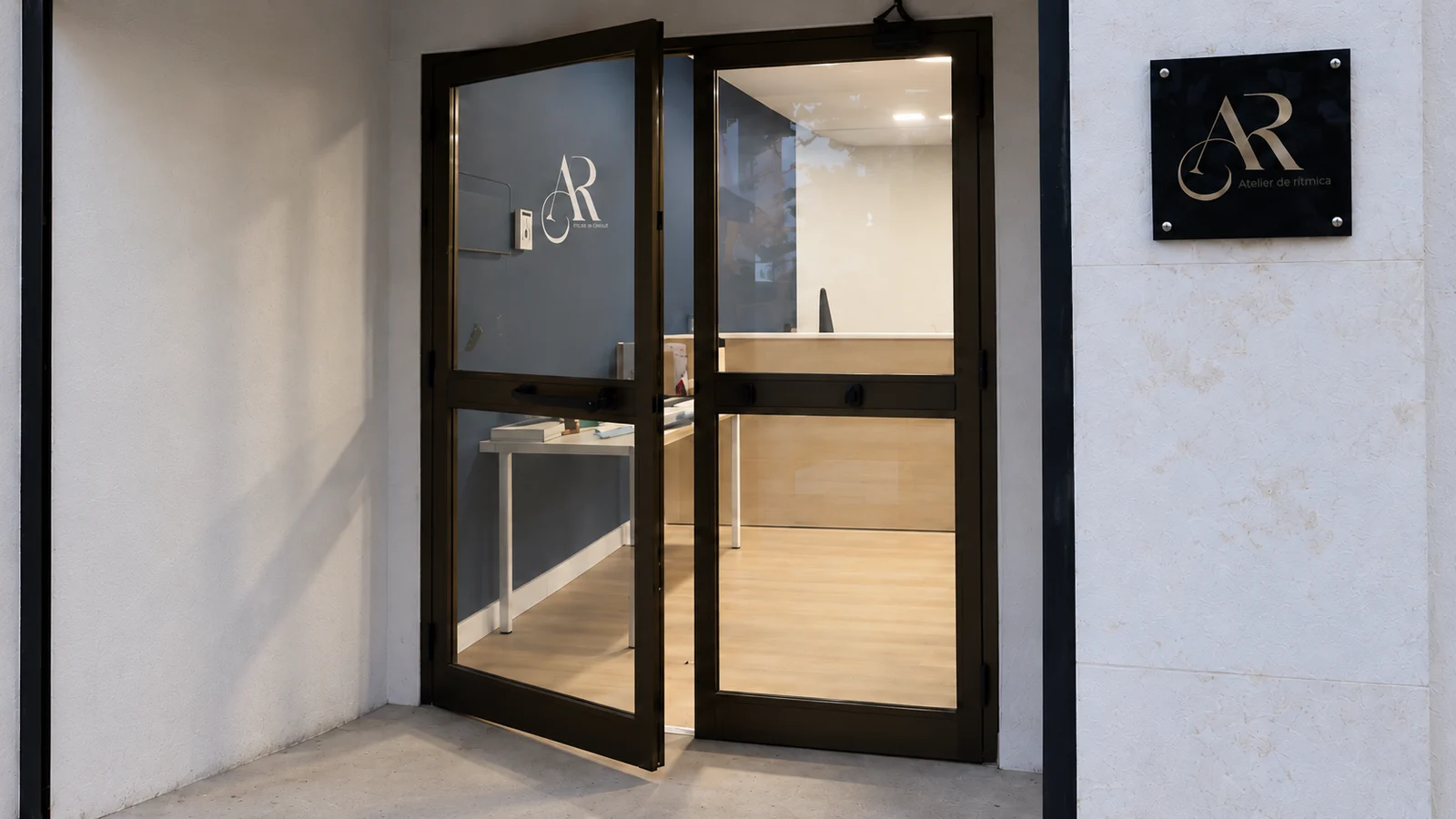 Puerta acristalada abierta con la placa dorada de Atelier de Rítmica en la jamba; al fondo se ve el mostrador de madera y la pared azul